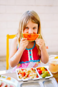 Picture of girl holding watermelon slice as a smile with a colorful lunchbox on the table in front of her