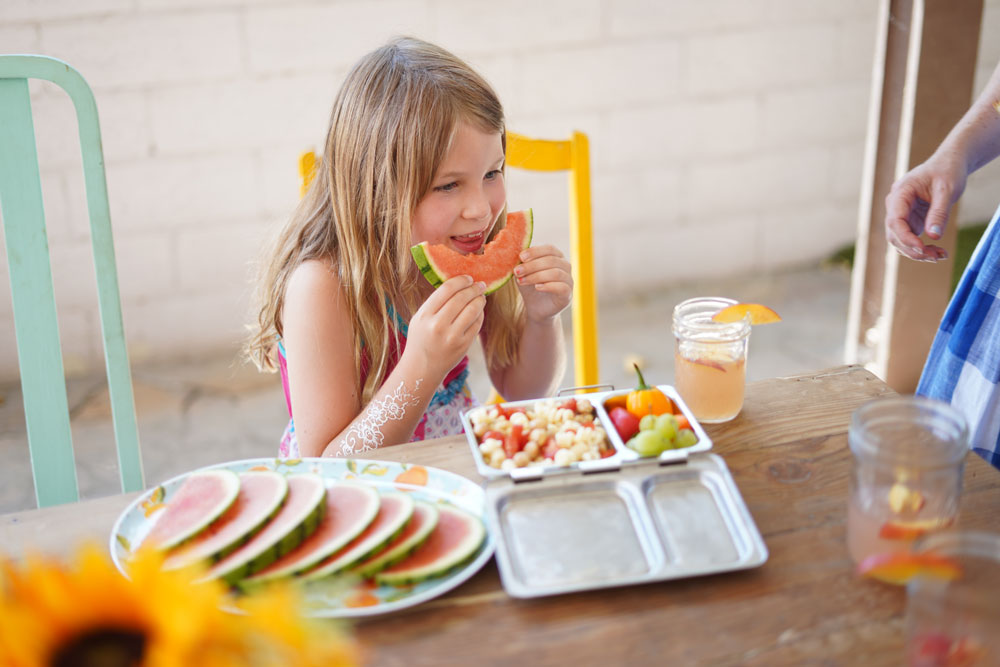 young girl eating a slice of watermelon with a variety of fruits and veggies in front of her