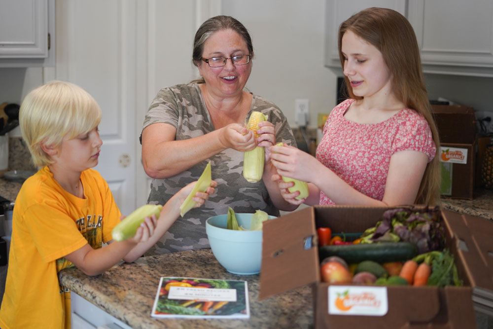 a mom and two kids shucking corn with a recipe book in front of them